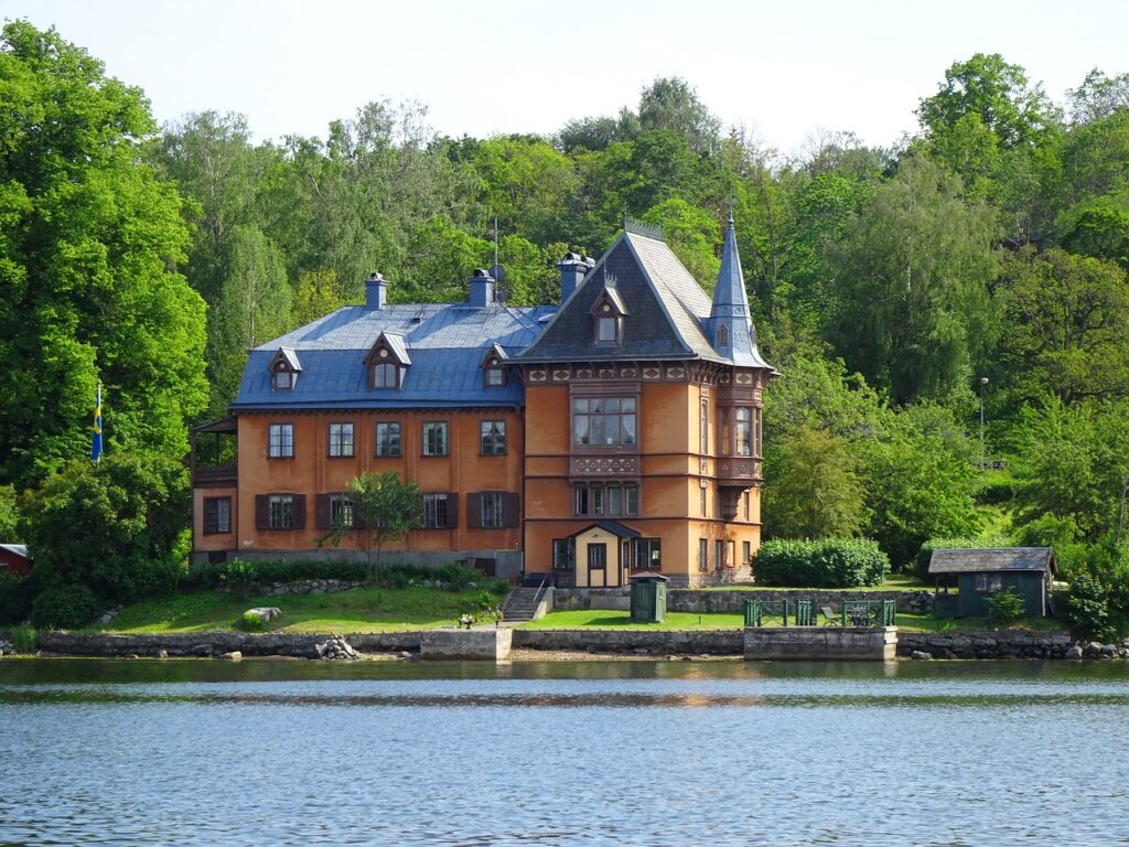 Large brick home with roofline that has windows, stone wall next to water