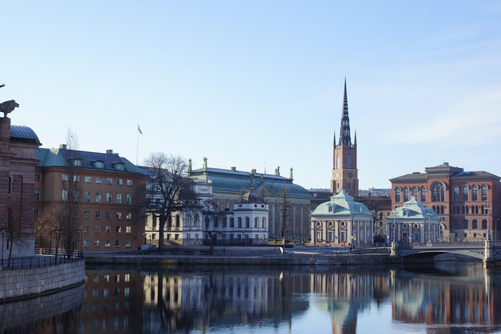 Buildings of various sizes on the edge of water with a bridge and a spire right of center.